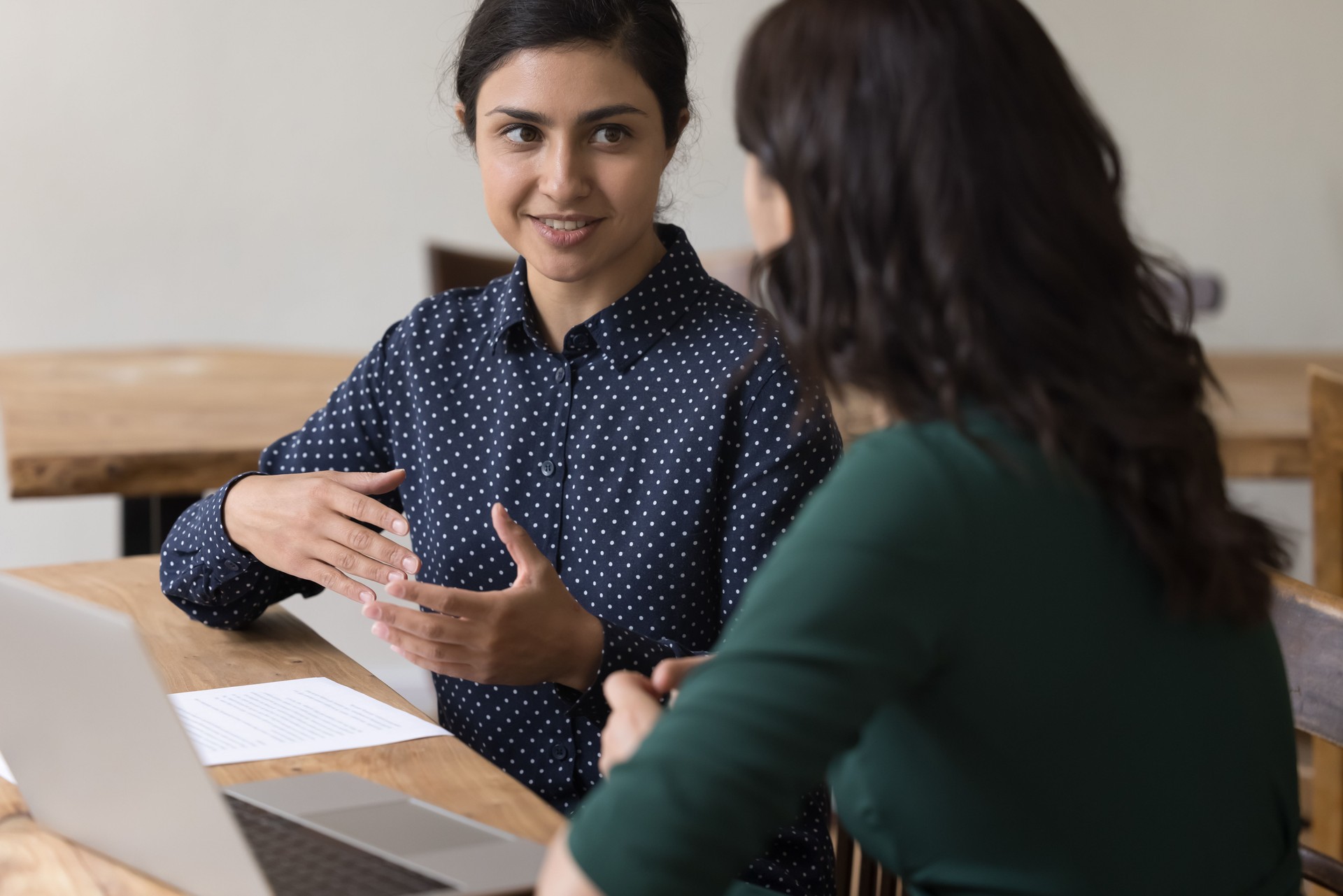 Women colleagues discuss joint task seated at desk with laptop